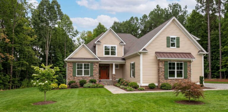 Front exterior of a two-story beige and stone house with green shutters, a brown front door, a metal awning over the front window, and a freshly landscaped lawn surrounded by mature forest trees.