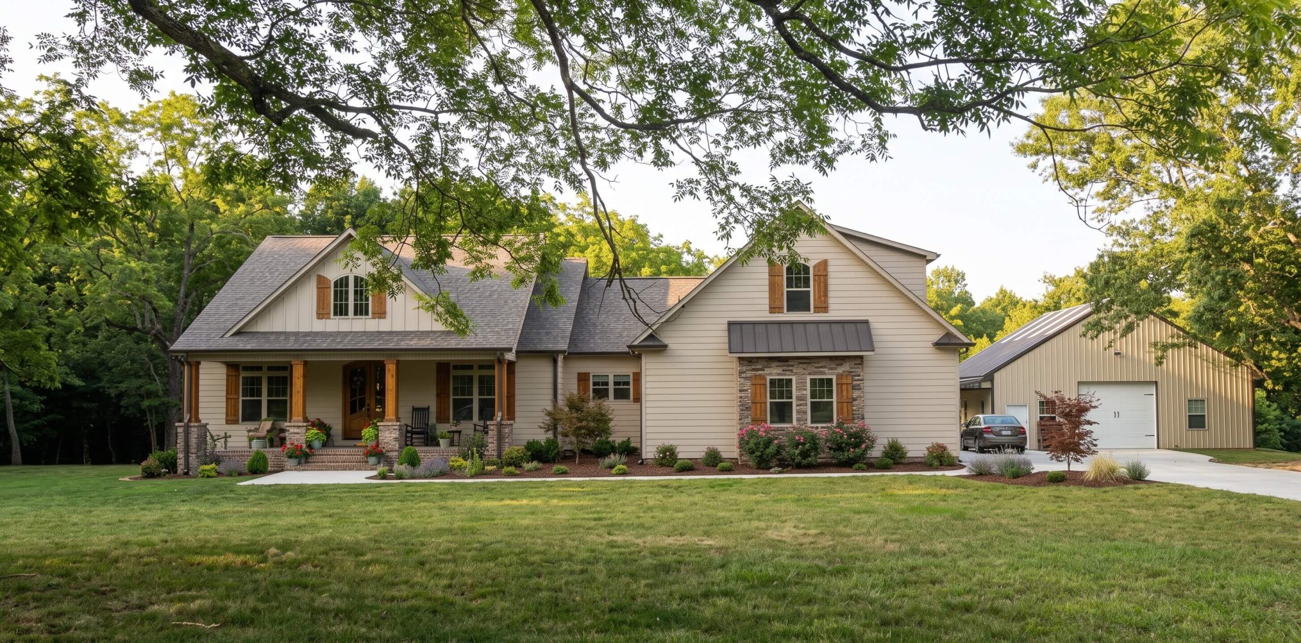 SEO-Optimized Alt Text: "Front view of a custom beige craftsman home with warm wood shutters, stone accents, a covered front porch, landscaped lawn, and a detached outbuilding garage on a wooded lot.